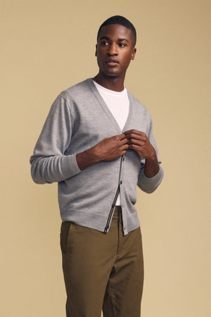A person stands against a beige background, wearing the SILVIO CARDIGAN by Velvet by Graham & Spencer—a premium wool cardigan with a V-neckline and ribbed trims—layered over a white shirt and olive green pants, hands adjusting the cardigan.