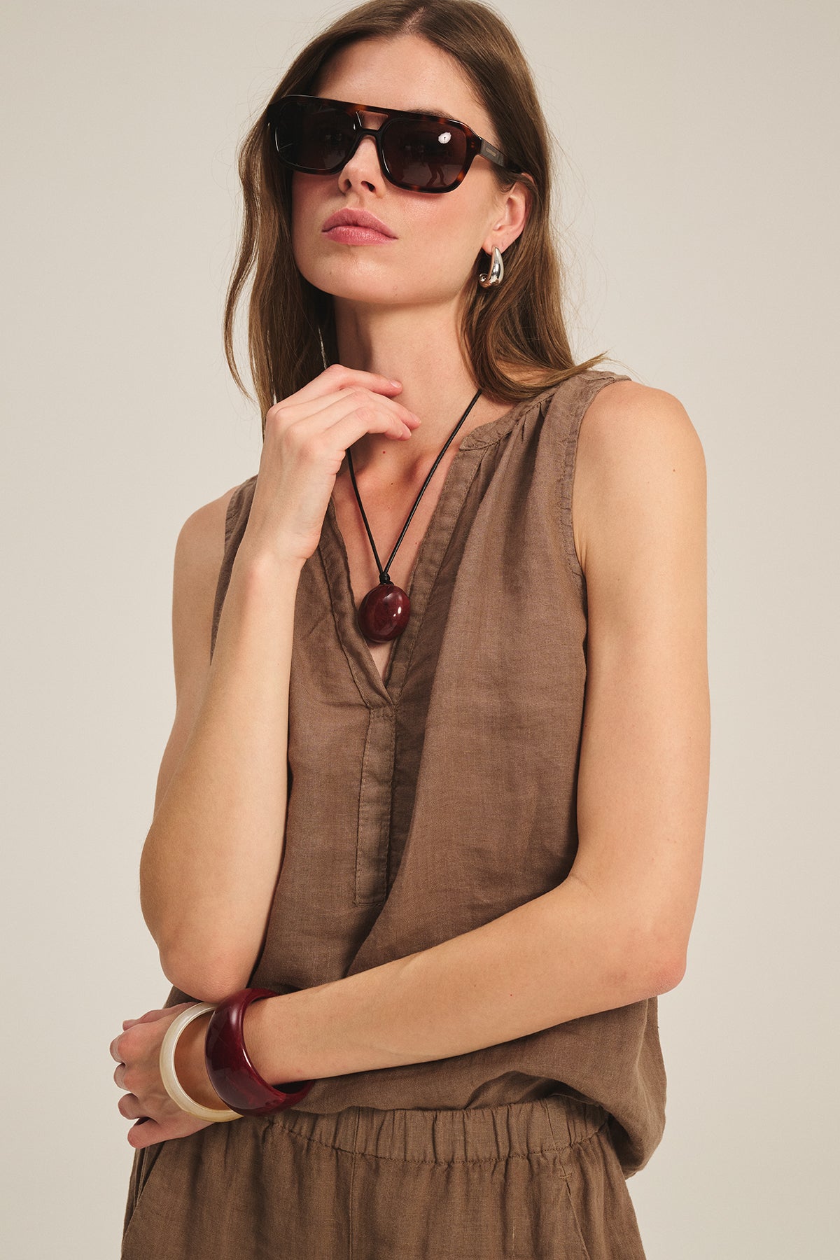 A woman wearing sunglasses and the TACY LINEN TANK TOP by Velvet by Graham & Spencer, featuring a brown v-neck sleeveless design and relaxed fit, paired with bold jewelry, stands against a neutral background.-39601053630657