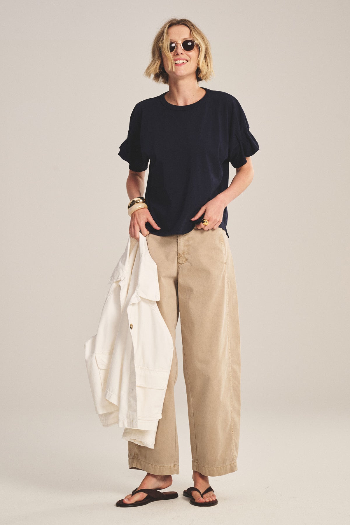 A woman in Velvet by Graham & Spencer's TAE TEE, a navy relaxed-fit crewneck top, pairs it with beige wide-leg pants, black sandals, sunglasses, and holds a white jacket against a plain background.-41008416948417