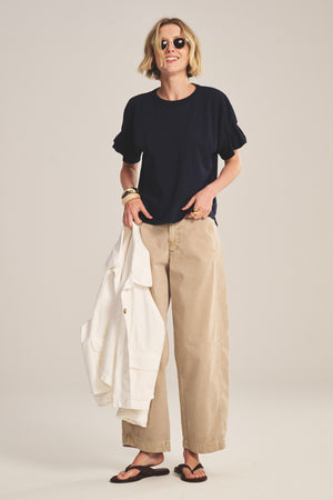 A woman in Velvet by Graham & Spencer's TAE TEE, a navy relaxed-fit crewneck top, pairs it with beige wide-leg pants, black sandals, sunglasses, and holds a white jacket against a plain background.