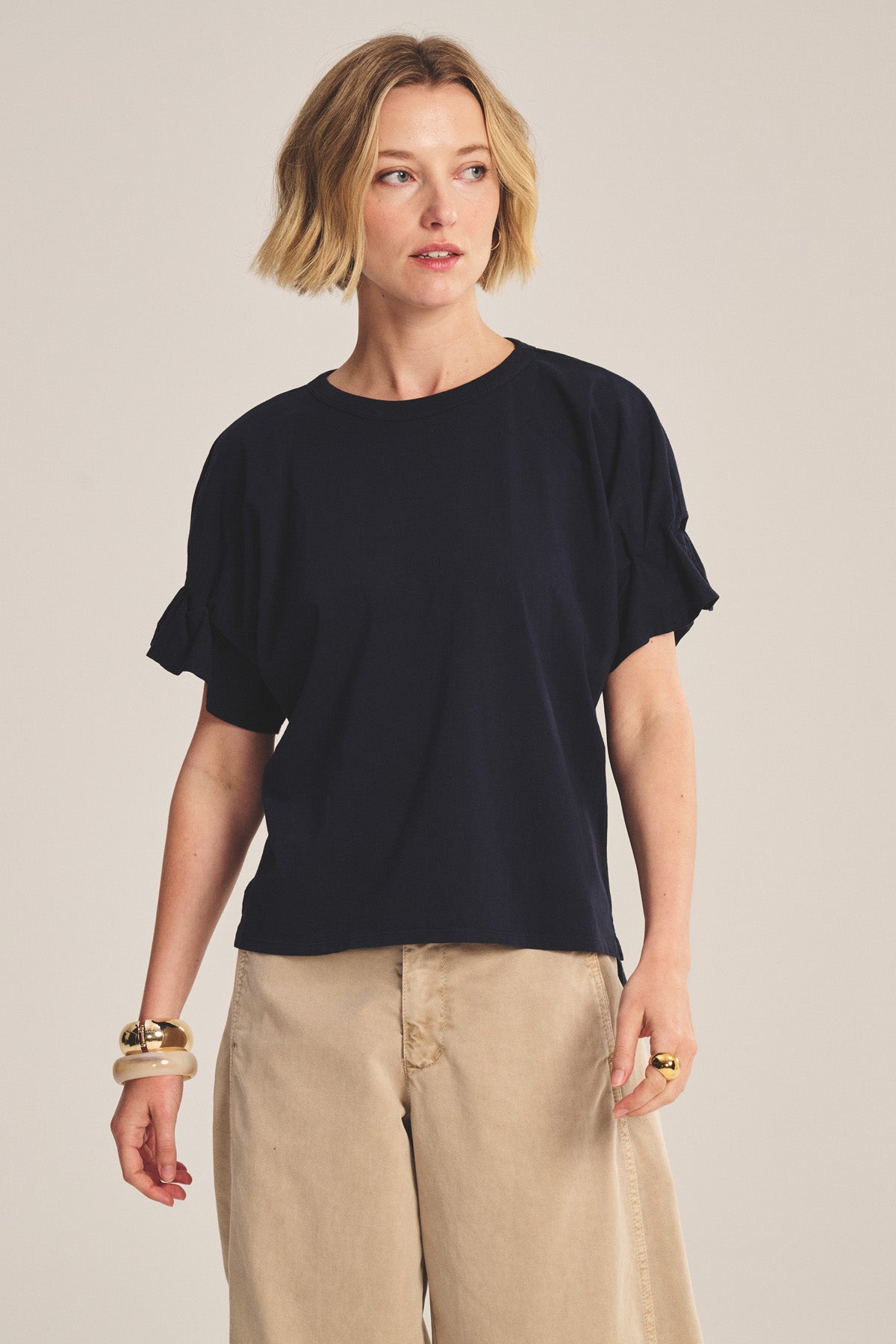 A woman with short blonde hair wears the TAE TEE by Velvet by Graham & Spencer—a navy cotton jersey relaxed-fit top with ruffled sleeves—paired with beige wide-leg pants and gold jewelry, against a light background.