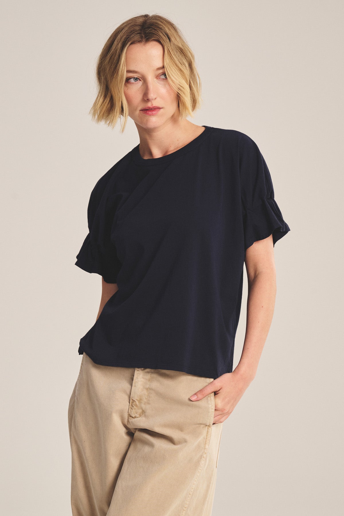 A woman with short blonde hair, wearing the Velvet by Graham & Spencer TAE TEE navy crewneck short-sleeve top and beige pants, stands with one hand in her pocket against a plain background.