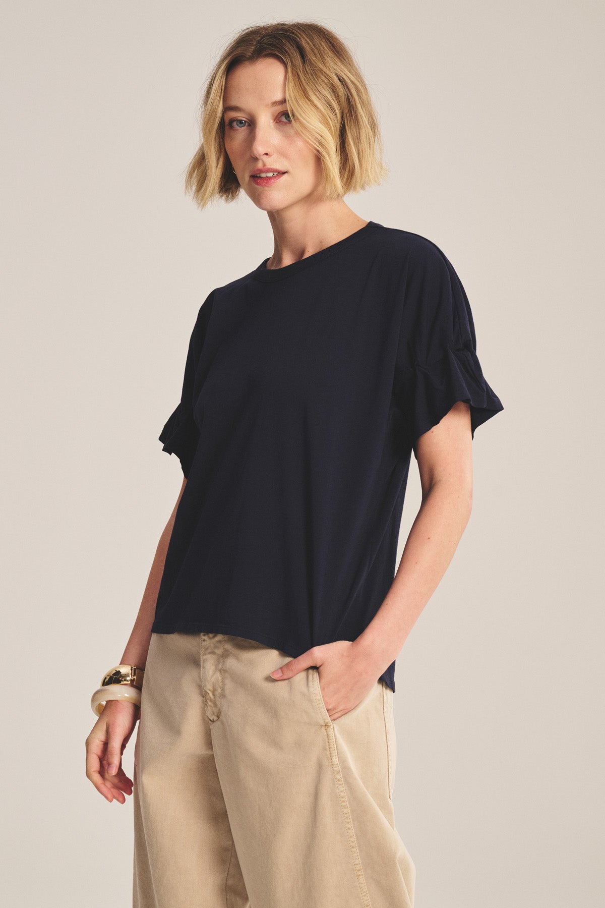 A woman with short blonde hair models the TAE TEE by Velvet by Graham & Spencer—a navy relaxed-fit crew neck tee with ruffled sleeves—paired with beige wide-leg pants against a neutral background.-41008416817345