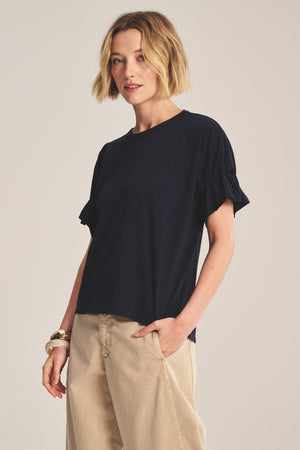 A woman with short blonde hair models the TAE TEE by Velvet by Graham & Spencer—a navy relaxed-fit crew neck tee with ruffled sleeves—paired with beige wide-leg pants against a neutral background.