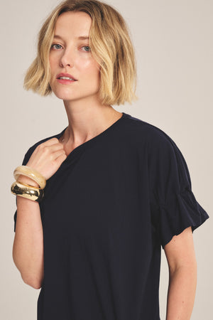 A woman with short blonde hair models the TAE TEE by Velvet by Graham & Spencer, a navy cotton jersey ruffled short-sleeve top, paired with large bangle bracelets against a plain background.