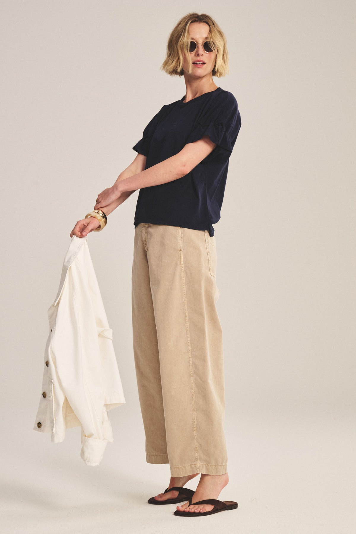 A woman models Velvet by Graham & Spencer’s TENLEY PANT—high-waisted beige cotton twill pants—with a navy top, black sandals, round sunglasses, and holds a white jacket in a studio setting.-41123276488897