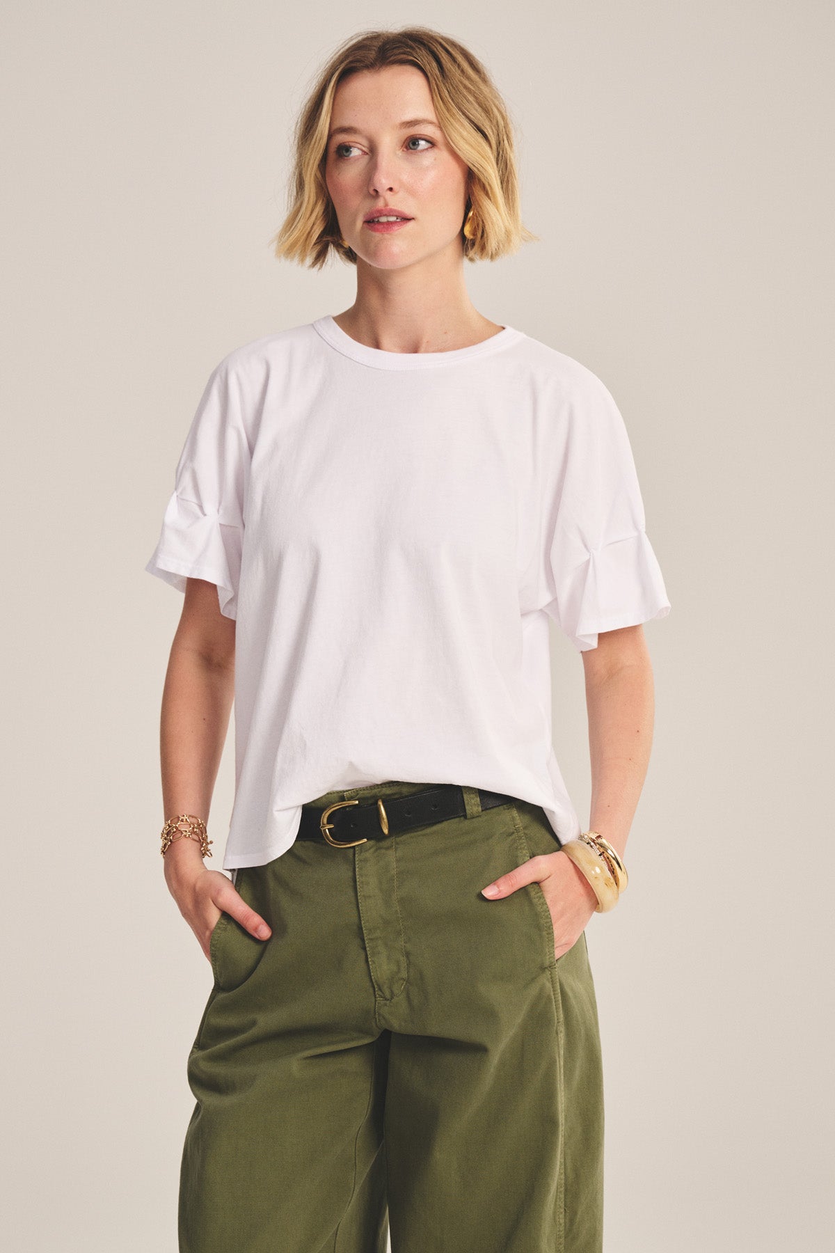 A woman with short blonde hair wears the TAE TEE from Velvet by Graham & Spencer in white cotton jersey, paired with olive green pants and a black belt, standing with her hands in her pockets.-40808728789185