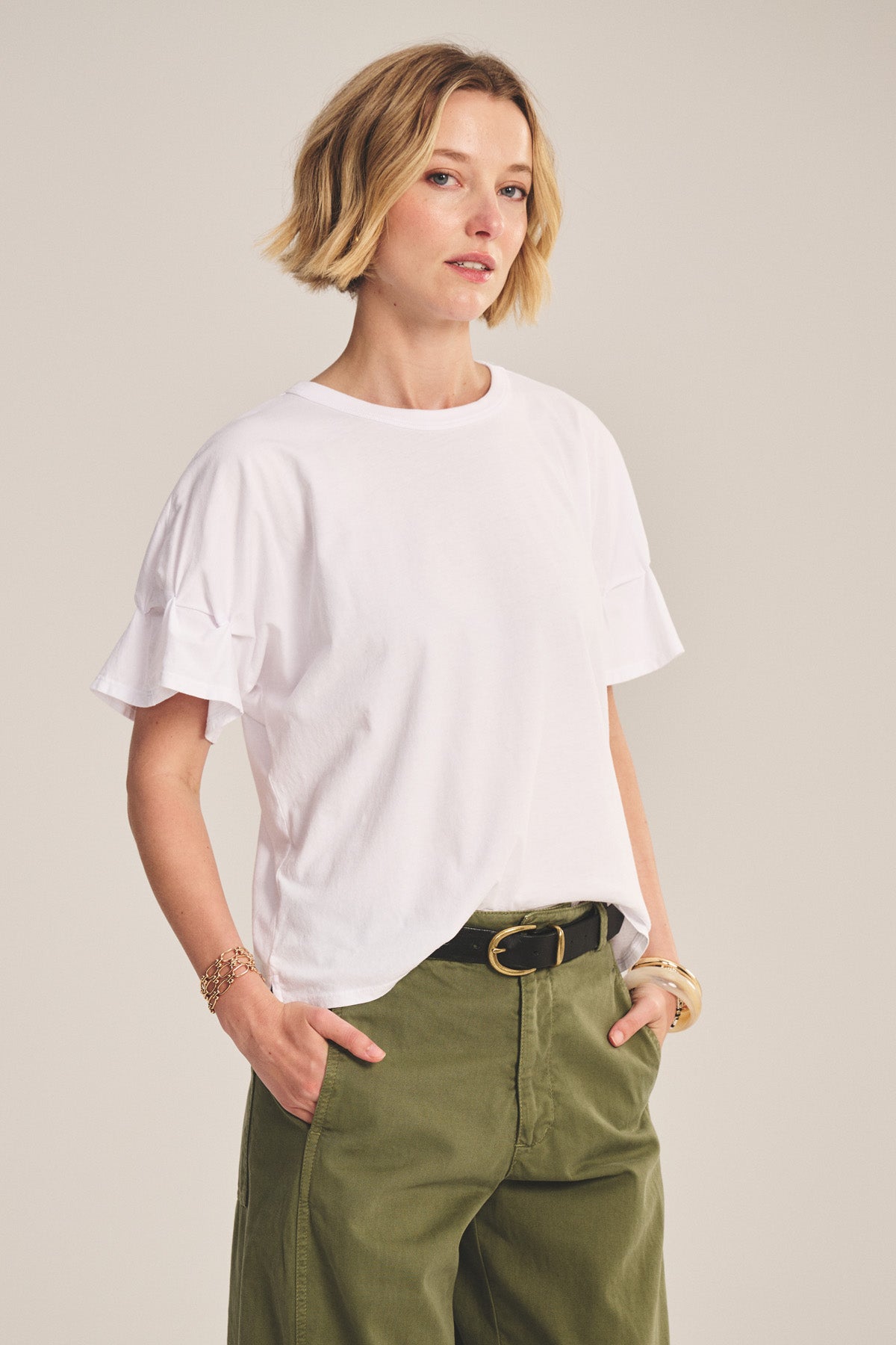 A woman with short blonde hair wears the TAE TEE by Velvet by Graham & Spencer in white cotton jersey, paired with olive green pants, hands in her pockets, posing against a plain background.-40808728756417