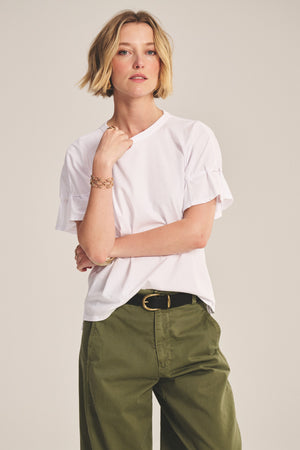A person with short blonde hair wears the TAE TEE by Velvet by Graham & Spencer—a white relaxed-fit top—paired with olive green pants, standing with one arm raised and the other hand on their waist.
