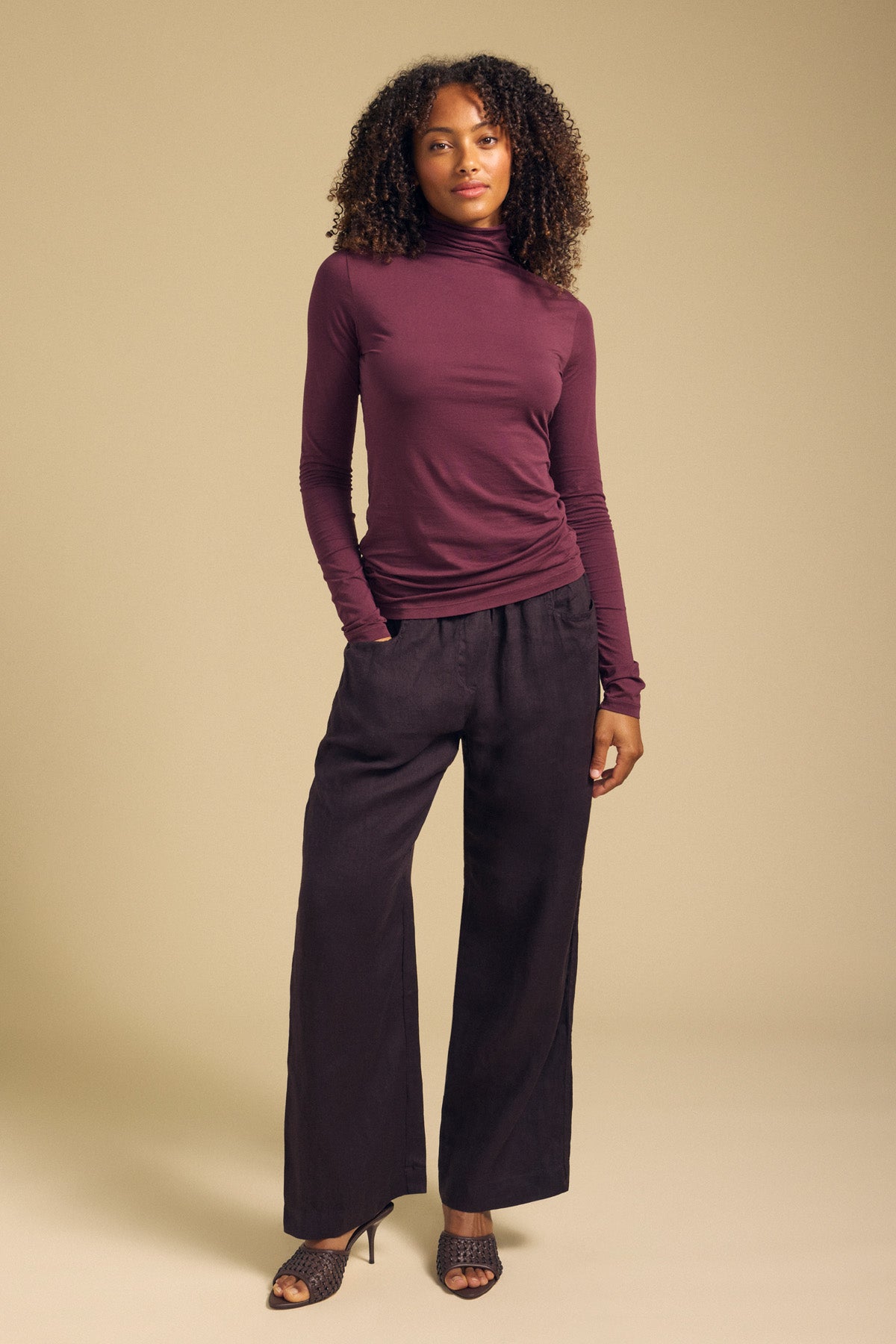 A person stands against a beige backdrop, wearing the Velvet by Graham & Spencer TALISIA TEE—a long-sleeve maroon mock neck top—paired with wide-leg black pants and black heeled sandals.