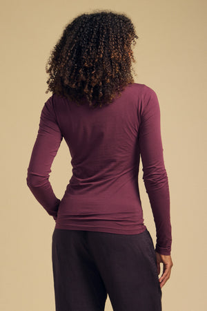 A person with curly hair is shown from the back, wearing the Velvet by Graham & Spencer TALISIA TEE—a long-sleeve maroon mock neck top and essential wardrobe staple—with dark pants, standing against a beige background.