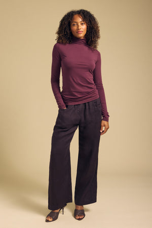 A person stands against a beige backdrop, wearing the Velvet by Graham & Spencer TALISIA TEE—a long-sleeve maroon mock neck top—paired with wide-leg black pants and black heeled sandals.