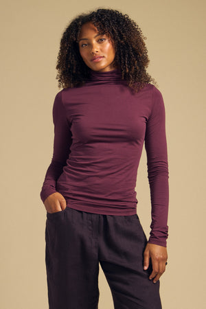 A woman wearing the Velvet by Graham & Spencer TALISIA TEE, a long-sleeve plum mock neck top, paired with dark pants stands with one hand in her pocket against a plain beige background.
