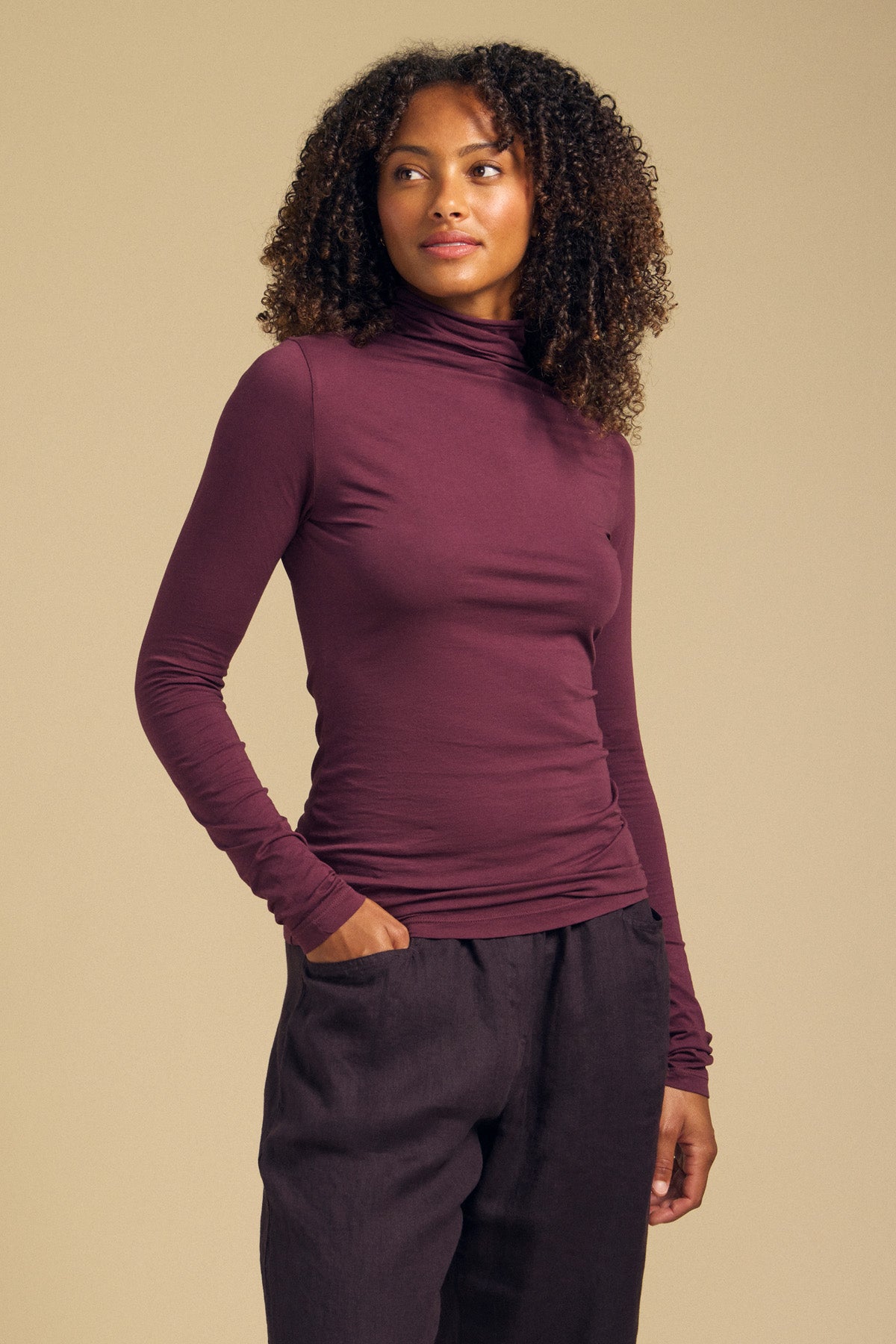A woman models the Velvet by Graham & Spencer TALISIA TEE—a long-sleeved, burgundy mock neck top—paired with dark pants, standing with one hand in her pocket against a plain beige background.-42063849947329