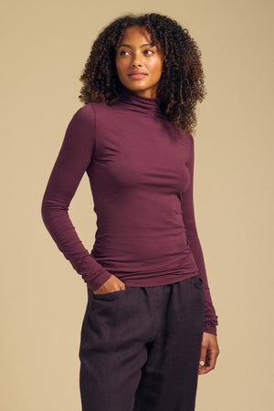 A woman models the Velvet by Graham & Spencer TALISIA TEE—a long-sleeved, burgundy mock neck top—paired with dark pants, standing with one hand in her pocket against a plain beige background.