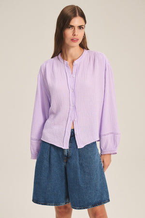A woman in the TARIA TOP by Velvet by Graham & Spencer—a light purple cotton gauze blouse with button-front closure and long sleeves—paired with blue knee-length denim shorts, stands against a plain background.