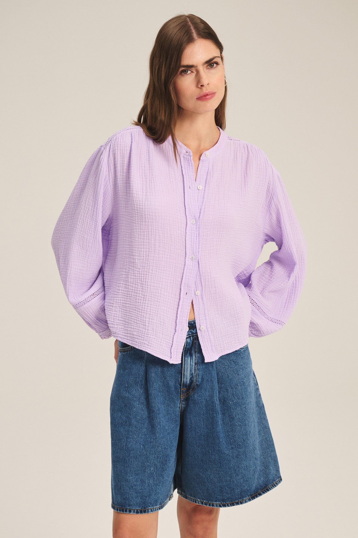 A woman wears the TARIA TOP by Velvet by Graham & Spencer, a light purple cotton gauze shirt with button-front closure, paired with knee-length blue denim shorts against a plain background.