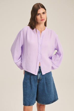 A woman wears the TARIA TOP by Velvet by Graham & Spencer, a light purple cotton gauze shirt with button-front closure, paired with knee-length blue denim shorts against a plain background.