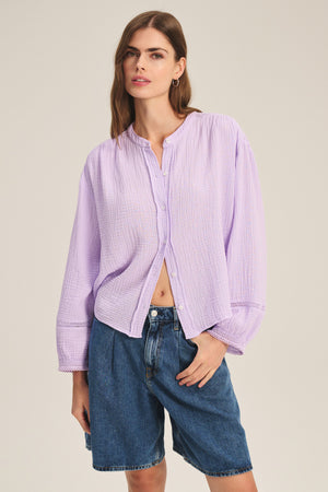 A woman wears the TARIA TOP by Velvet by Graham & Spencer, a light purple cotton gauze button-front blouse with long sleeves, paired with blue knee-length denim shorts, standing against a plain background.