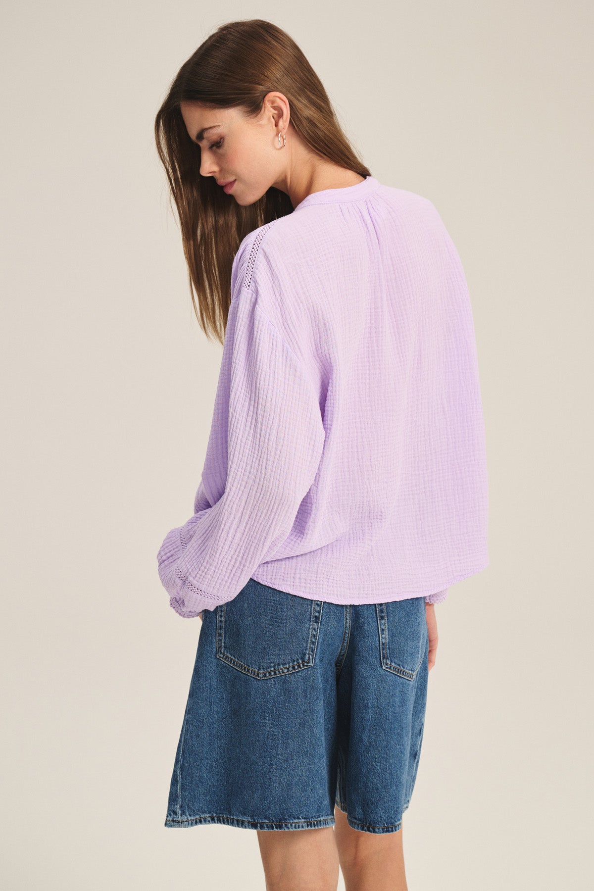 Wearing the Velvet by Graham & Spencer TARIA TOP—a loose, long-sleeve cotton gauze shirt with button-front closure—and knee-length blue denim shorts, a person stands sideways to the camera, looking down.-41417904160961
