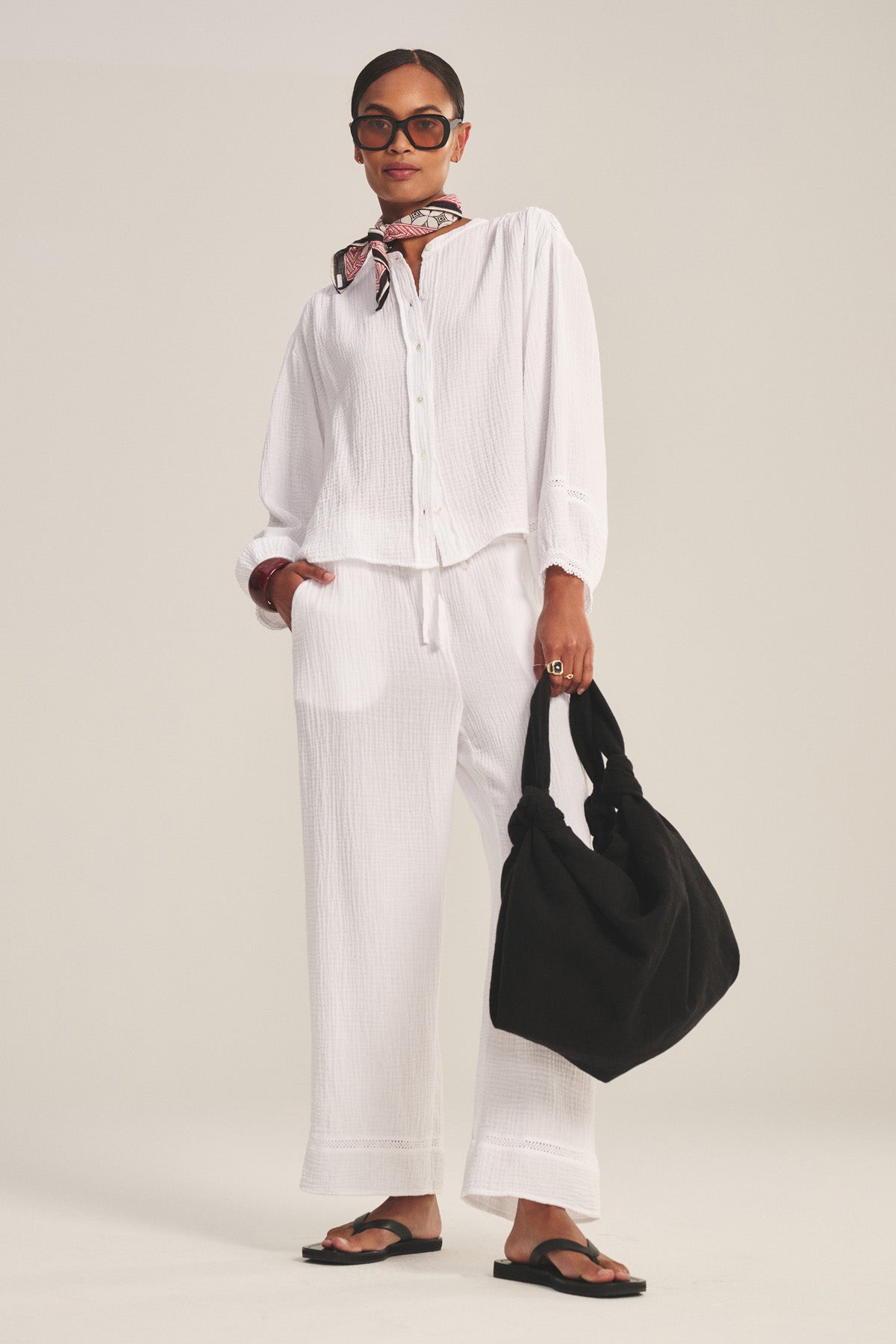 A woman in the TARIA TOP by Velvet by Graham & Spencer, a white cotton gauze button-front shirt, styled with matching pants, black sandals, a neck scarf, sunglasses, and carrying a black bag.-41128866840769