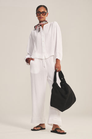 A woman in the TARIA TOP by Velvet by Graham & Spencer, a white cotton gauze button-front shirt, styled with matching pants, black sandals, a neck scarf, sunglasses, and carrying a black bag.