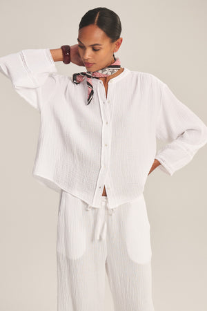 A person wears the TARIA TOP by Velvet by Graham & Spencer with matching white pants and a red patterned scarf, standing against a plain background for an effortlessly lightweight and comfortable look.