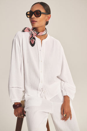 Wearing large sunglasses, a patterned neck scarf, the TARIA TOP by Velvet by Graham & Spencer—a loose cotton gauze button-front blouse—and white pants, a person sits on a stool against a plain background.
