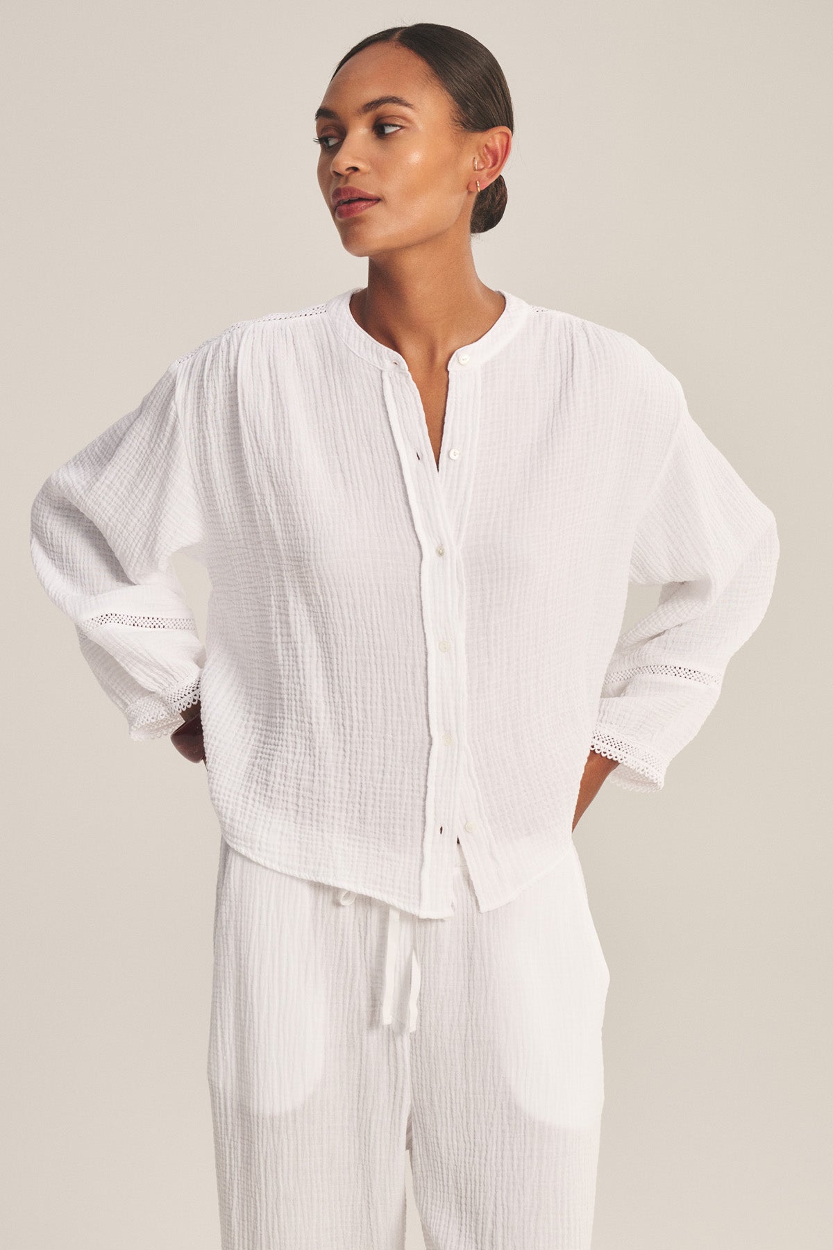 A woman stands with hands on hips against a plain background, modeling the TARIA TOP by Velvet by Graham & Spencer—a loose-fitting cotton gauze shirt with button-front closure, paired with matching white pants.-41128866709697