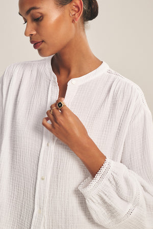 A woman wears the TARIA TOP by Velvet by Graham & Spencer, a white textured cotton gauze blouse with lace-trimmed cuffs and a button-front closure. She looks down, touching her collar, accessorized with gold hoops and a black gemstone ring.