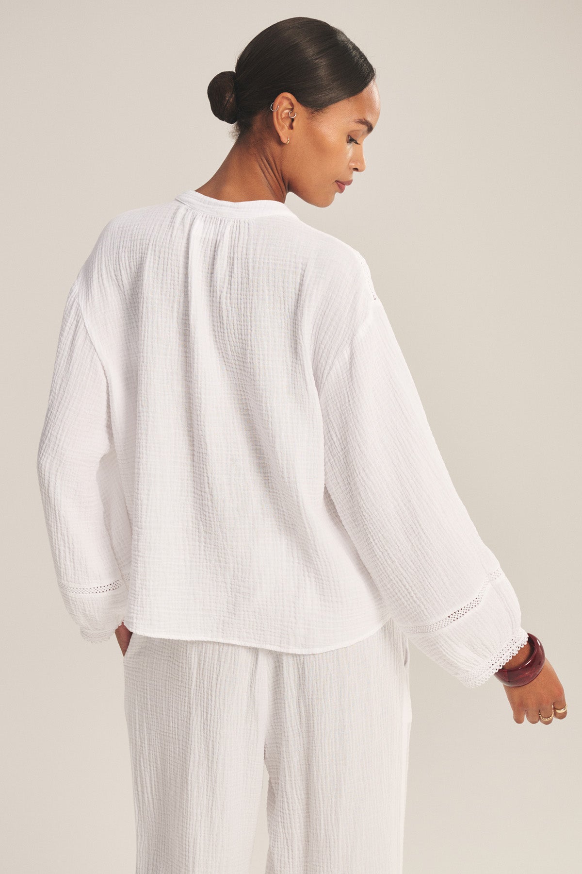 A woman stands with her back to the camera, wearing the TARIA TOP by Velvet by Graham & Spencer—a loose-fitting white cotton gauze top paired with matching pants. Her hair is in a bun, set against a plain background.