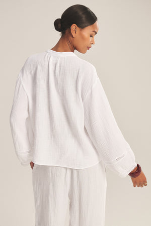 A woman stands with her back to the camera, wearing the TARIA TOP by Velvet by Graham & Spencer—a loose-fitting white cotton gauze top paired with matching pants. Her hair is in a bun, set against a plain background.