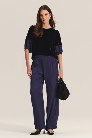 A woman poses against a light background wearing Velvet by Graham & Spencer’s LARKIN PANT—wide-leg satin pants styled with a dark velvet top, black bag, and black heels, creating a flattering everyday look.