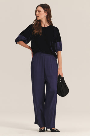 A woman wears Velvet by Graham & Spencer’s LARKIN PANT, a wide-leg satin style in navy, paired with a matching three-quarter sleeve velvet top. She accessorizes with a small black bag and embellished heels for an elevated everyday look.