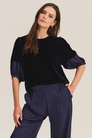 A woman stands with one hand on her hip, modeling the LARKIN PANT from Velvet by Graham & Spencer—wide-leg satin pants with a flattering fit, ideal for everyday wear—paired with a dark velvet top against a plain background.