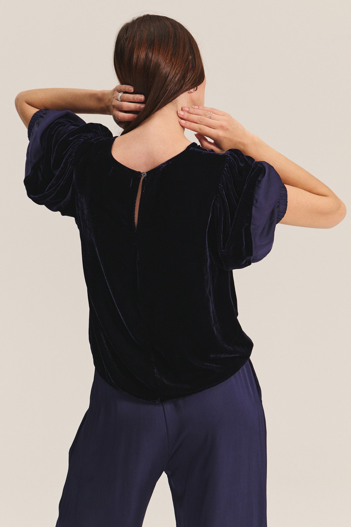 A person with brown hair in a low ponytail is seen from the back, wearing the LARKIN PANT by Velvet by Graham & Spencer—wide-leg satin pants styled with a navy velvet puff-sleeve top—for a flattering, everyday look.-40263652868289