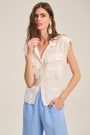 A woman wears the Velvet by Graham & Spencer TASHA LINEN TOP, a sleeveless lightweight cargo style with two chest pockets, paired with light blue wide-leg pants, standing against a plain background.