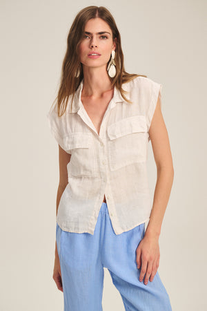 A woman wears the TASHA LINEN TOP by Velvet by Graham & Spencer—a sleeveless, breathable linen top with front pockets—paired with light blue pants, standing against a neutral background.