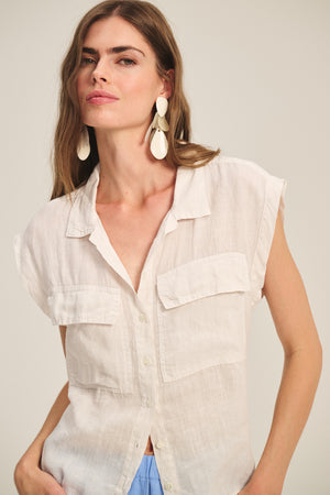 A woman wears the TASHA LINEN TOP by Velvet by Graham & Spencer, a light beige, breathable linen shirt with chest pockets, paired with blue pants and large white earrings, standing against a plain background.