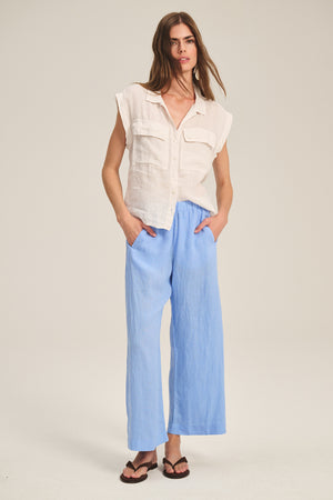 A woman stands against a plain background, wearing the TASHA LINEN TOP by Velvet by Graham & Spencer, paired with light blue wide-leg pants and black sandals, her hands casually in her pockets.