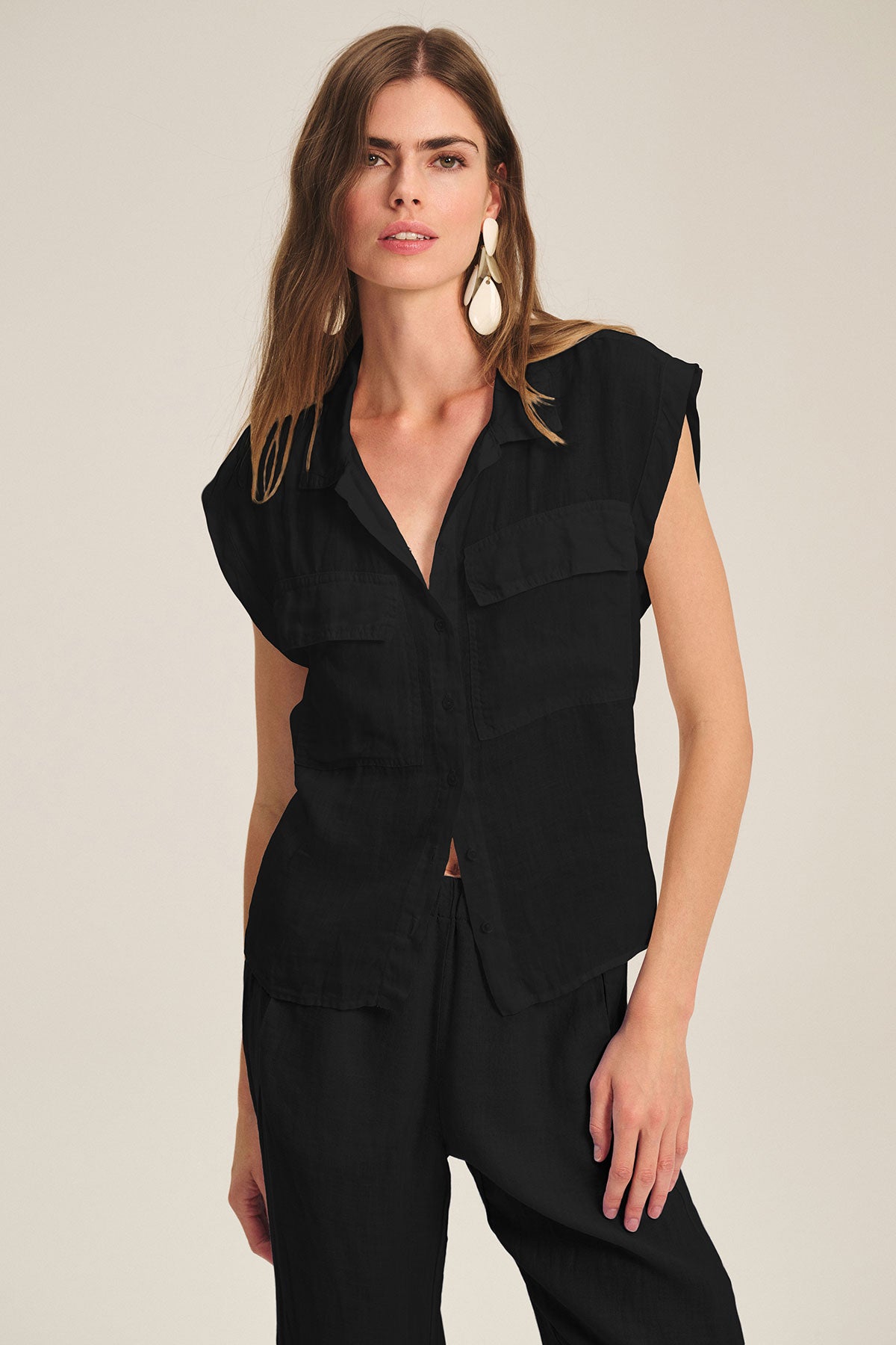   A woman wears the TASHA LINEN TOP by Velvet by Graham & Spencer—sleeveless, with chest pockets—paired with matching black pants against a plain background. 