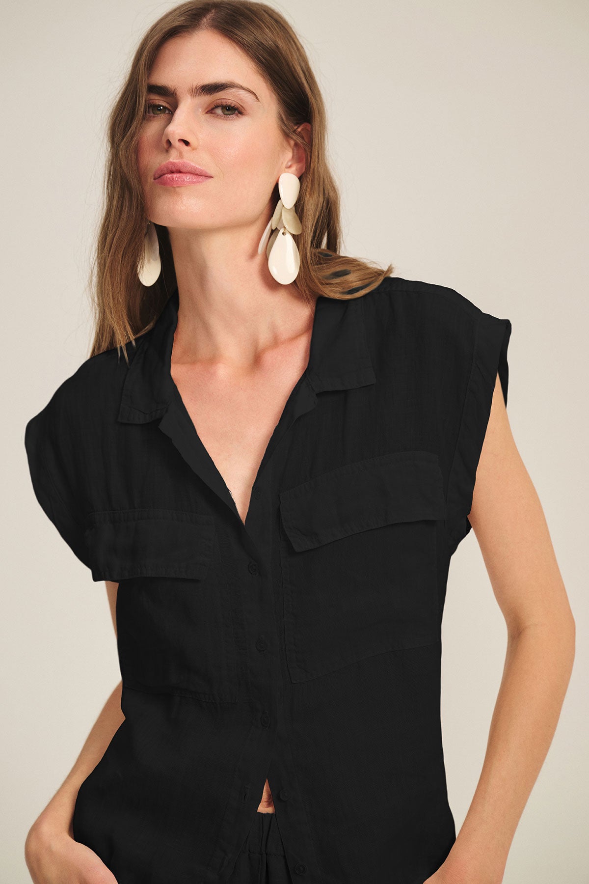 A woman poses against a plain background wearing the TASHA LINEN TOP by Velvet by Graham & Spencer, a black linen cargo style featuring two chest pockets, styled with large white dangling earrings.-39725079593153