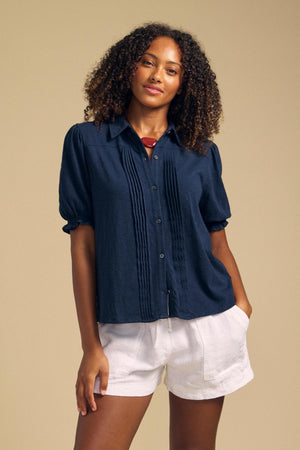 A woman stands with one hand in her pocket against a plain beige background, wearing the TATUM TEE by Velvet by Graham & Spencer—a navy feminine blouse with pleats—paired with white shorts.