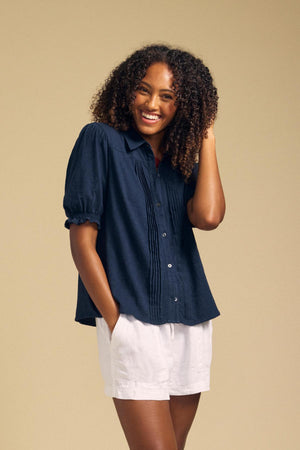 Wearing the TATUM TEE by Velvet by Graham & Spencer, a person with curly hair pairs the navy button front top with white shorts, standing against a beige background and smiling as they touch their hair.