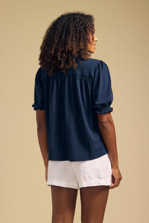 A person stands facing away against a plain beige background, wearing the TATUM TEE by Velvet by Graham & Spencer—a navy blue puffed sleeve blouse with gathered details—paired with white shorts.