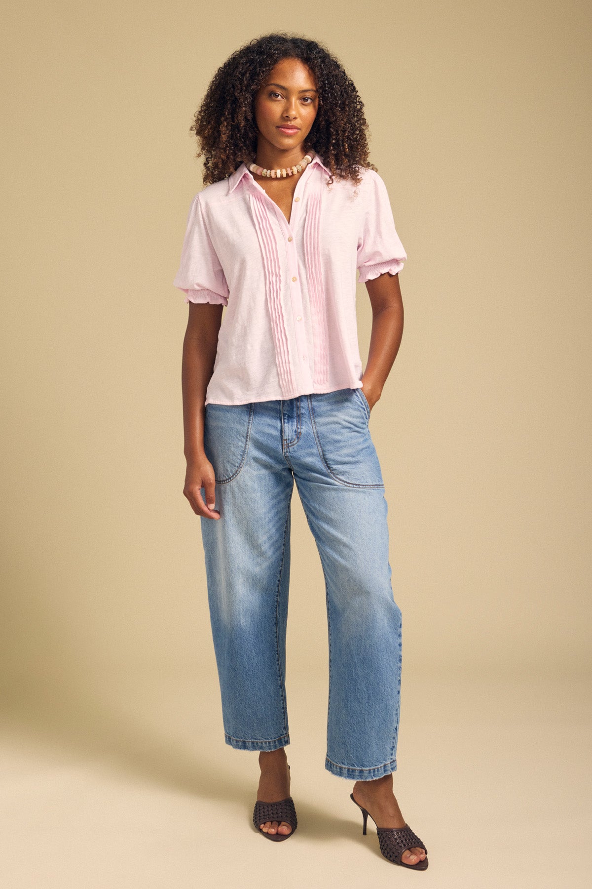 A person stands against a beige background wearing the Velvet by Graham & Spencer TATUM TEE—a light pink cotton slub tee featuring subtle puffed sleeves—paired with blue wide-leg jeans, dark heeled sandals, and a pearl necklace.-41934525661377