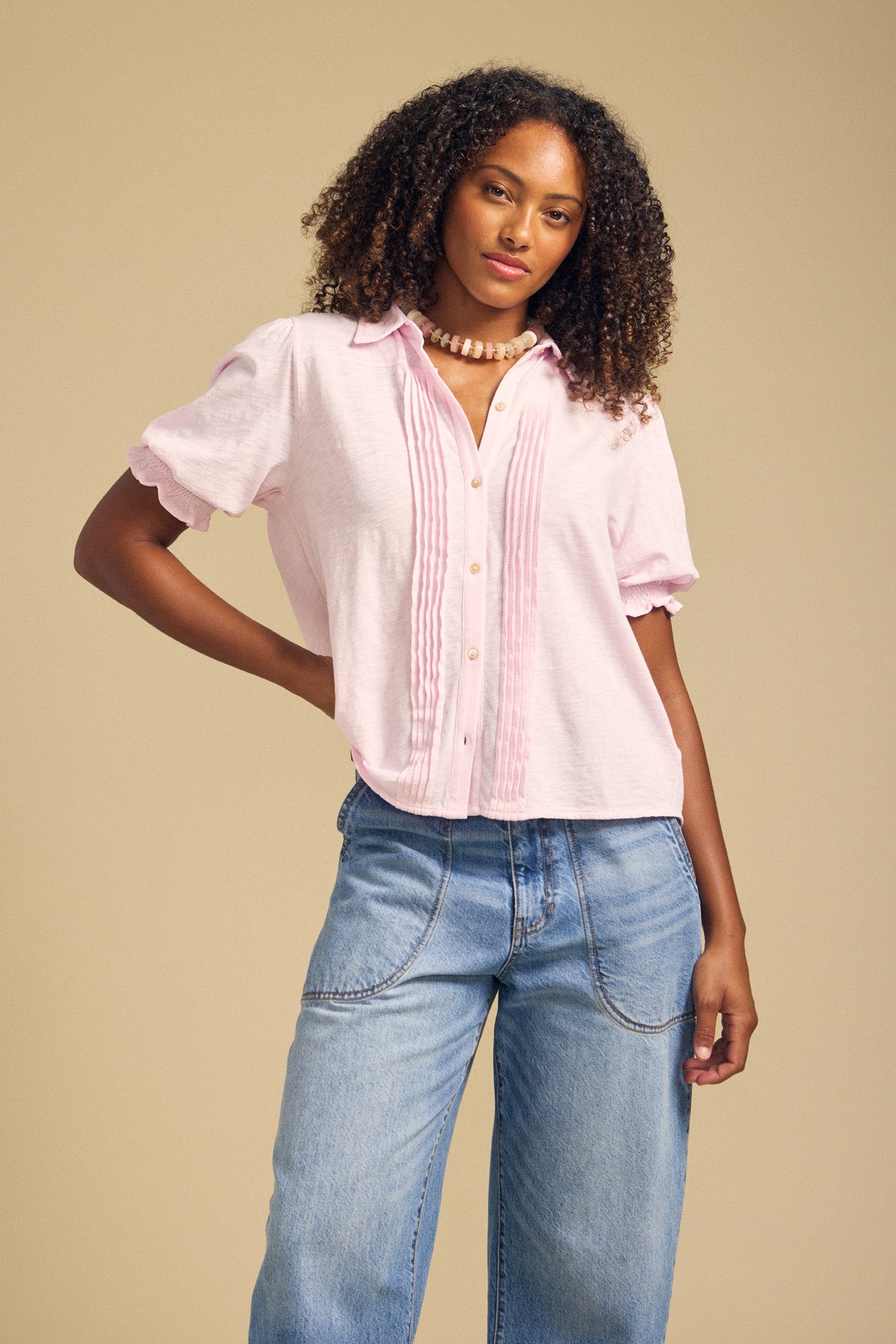 A person wearing the TATUM TEE by Velvet by Graham & Spencer—a light pink feminine blouse with pintuck details—and light blue wide-leg jeans stands against a plain beige background.-41934525628609