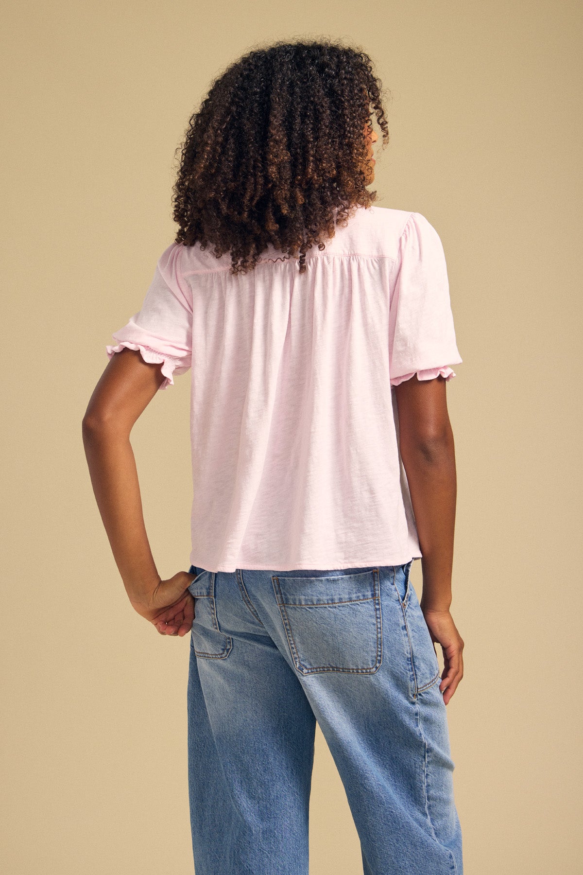 A person stands with their back to the camera against a plain beige background, wearing Velvet by Graham & Spencer’s TATUM TEE in light pink cotton slub with ruffled edges, paired with blue jeans.
