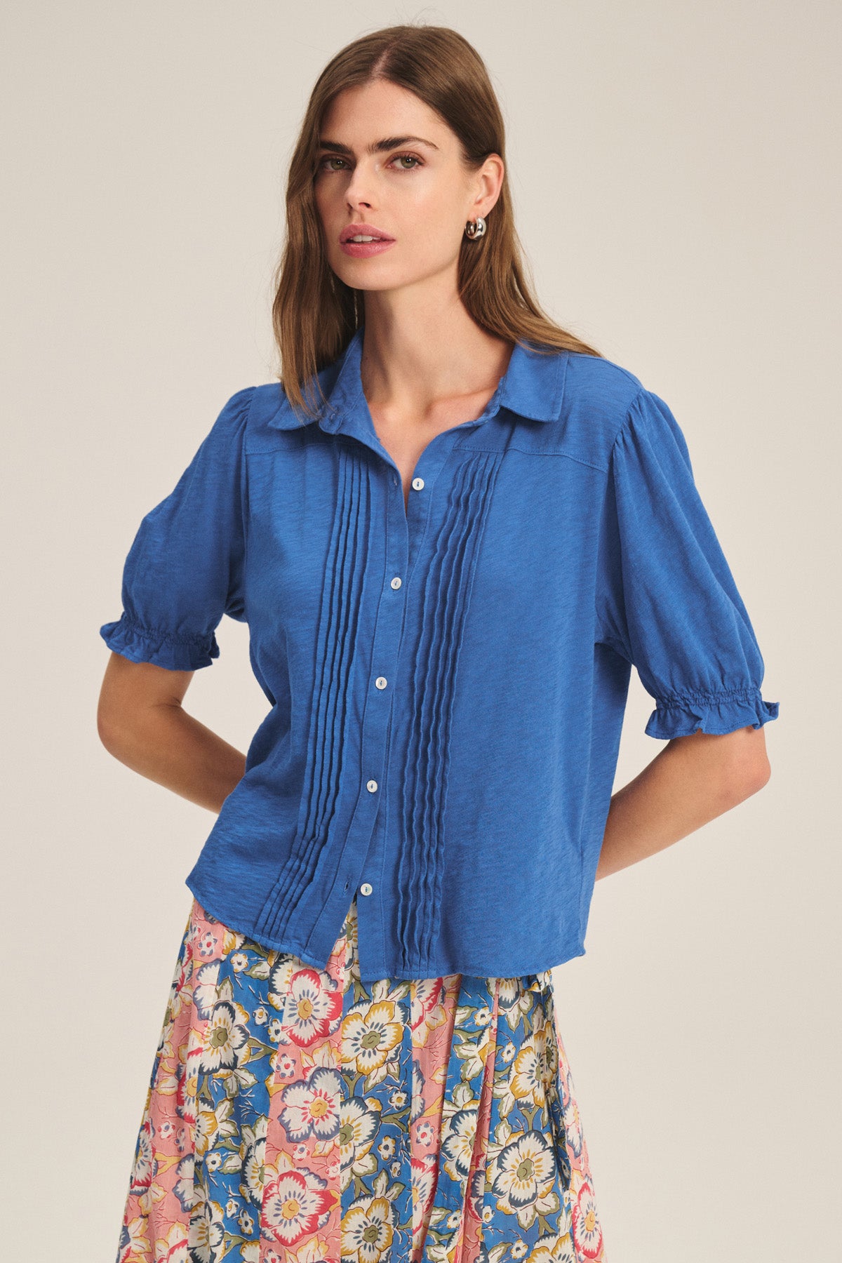A woman stands against a plain background wearing the Velvet by Graham & Spencer TATUM TEE, a blue button-front top with pintuck detailing, paired with a floral skirt.