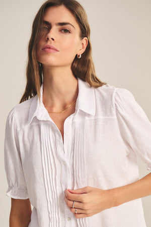 A woman wears the TATUM TEE by Velvet by Graham & Spencer—a white, button-front blouse with pleated details and subtle puff sleeves—standing against a neutral background.